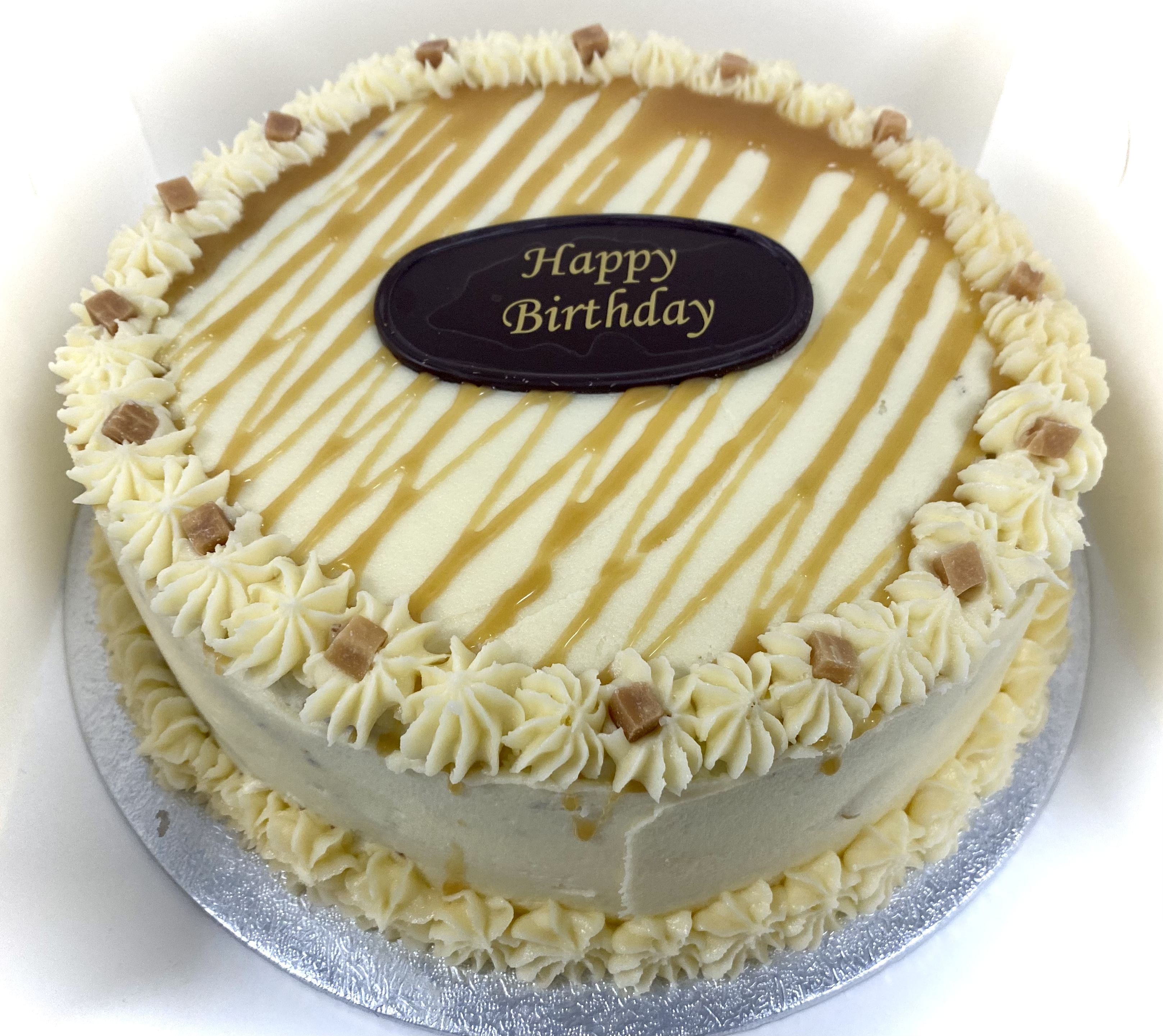 A round, vanilla gateaux frosted cake decorated with caramel drizzle and small cream rosettes around the edge. Topped with a chocolate plaque that reads 'Happy Birthday' in yellow lettering. The cake, infused with salted caramel frosting, sits on a silver cake board.